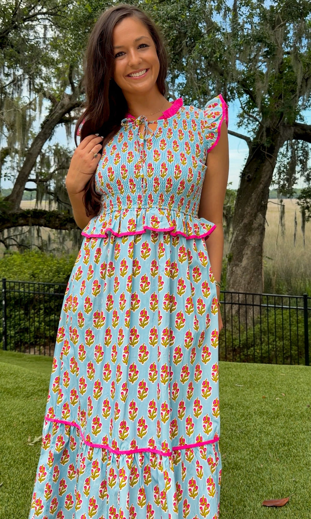 Light blue floral print midi dress featuring a fully smocked bodice, flutter sleeves with pink contrast trim, a keyhole tie neckline, ruffled peplum waist, tiered midi skirt, and side pockets.