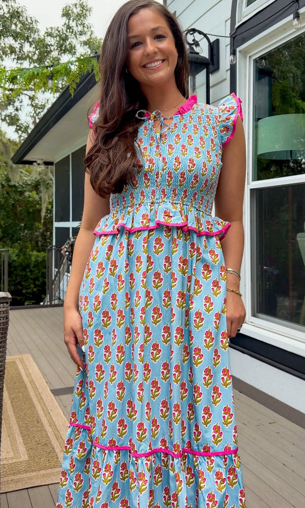 A woman wearing a light blue maxi dress with a colorful floral print and bright pink ruffle trim, standing on a wooden deck in front of a house.