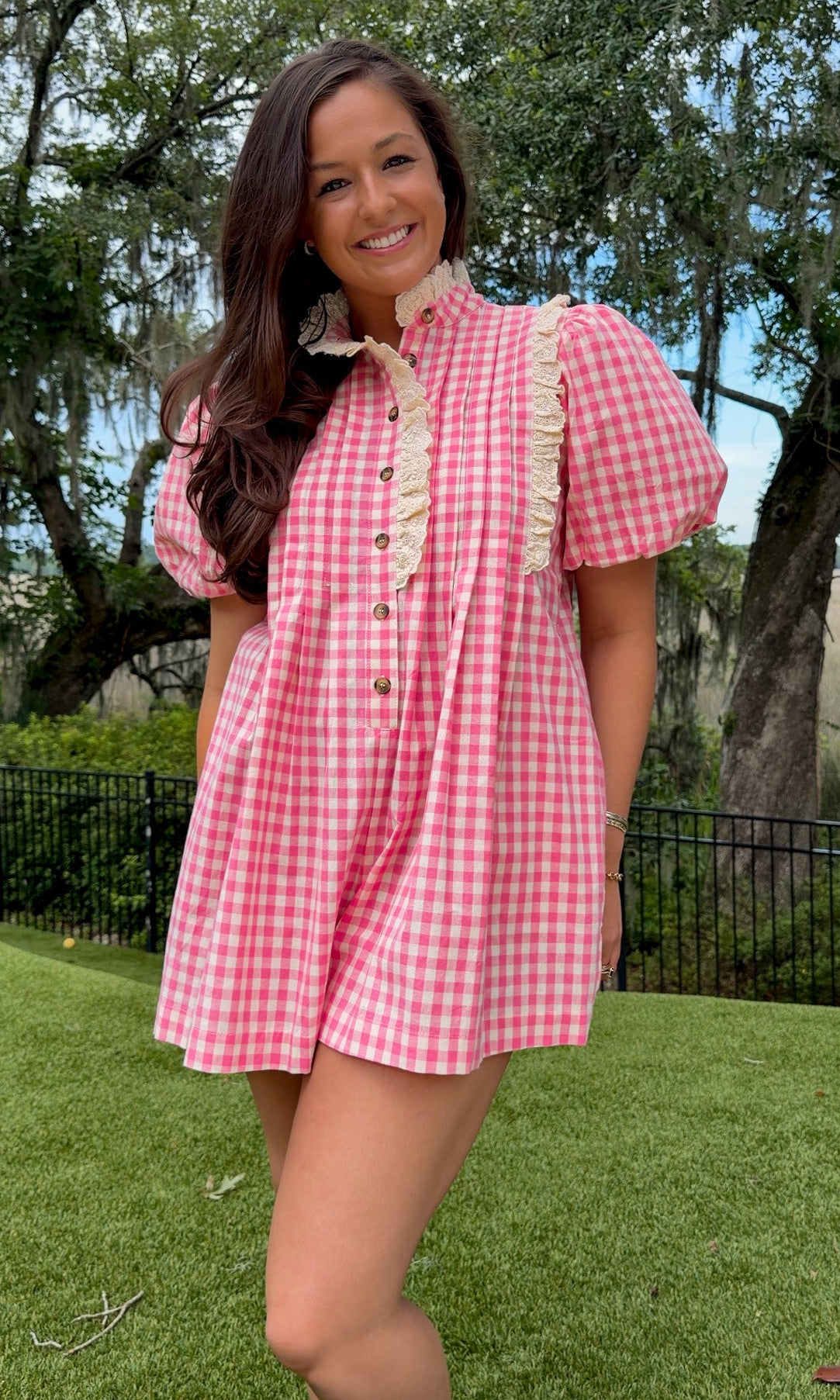 A woman wearing a pink and white gingham short-sleeve mini dress with cream ruffle trim and buttons, standing outdoors on a green lawn with trees in the background.