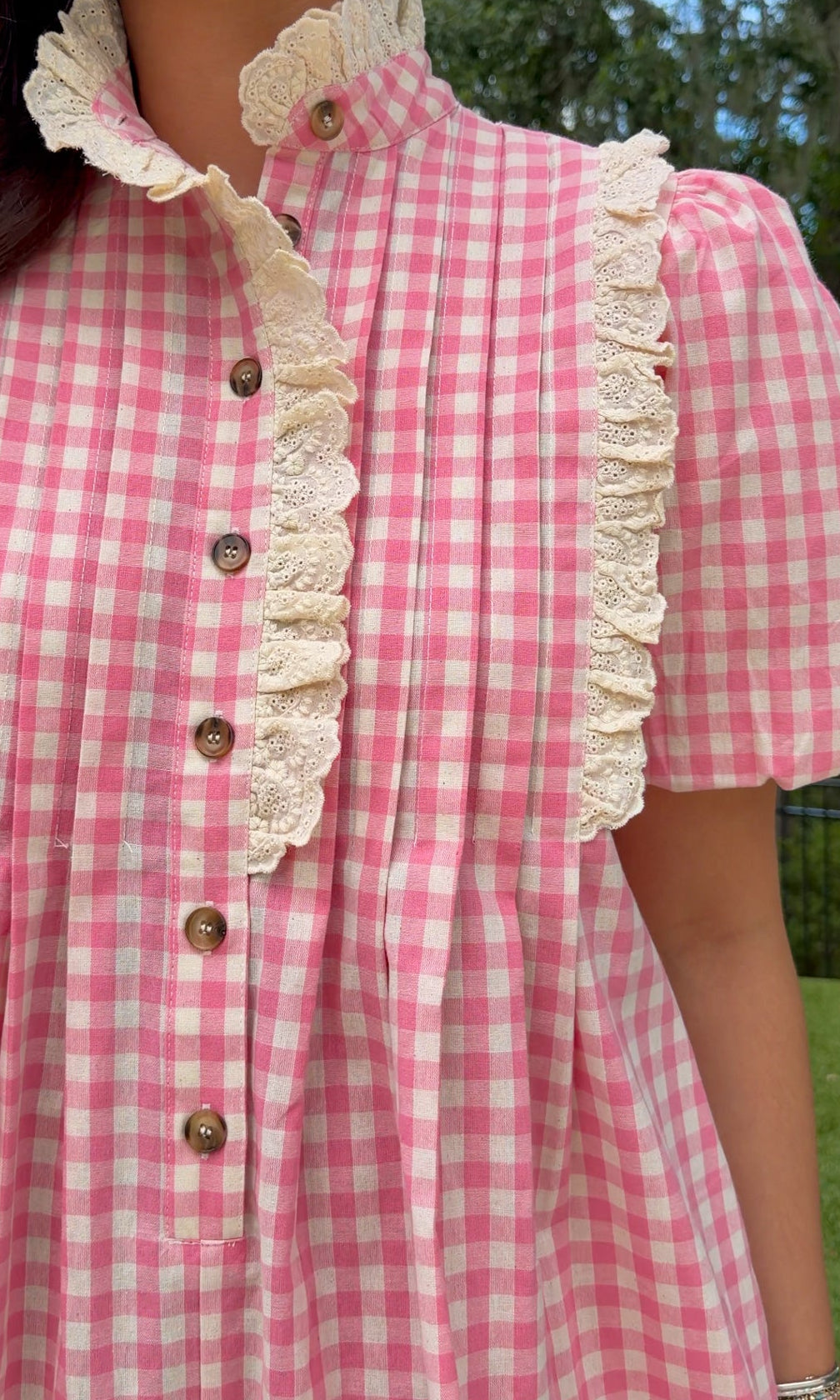A close-up of a person wearing a pink and white gingham dress with cream lace trim and brown buttons, photographed outdoors with trees and grass in the background.