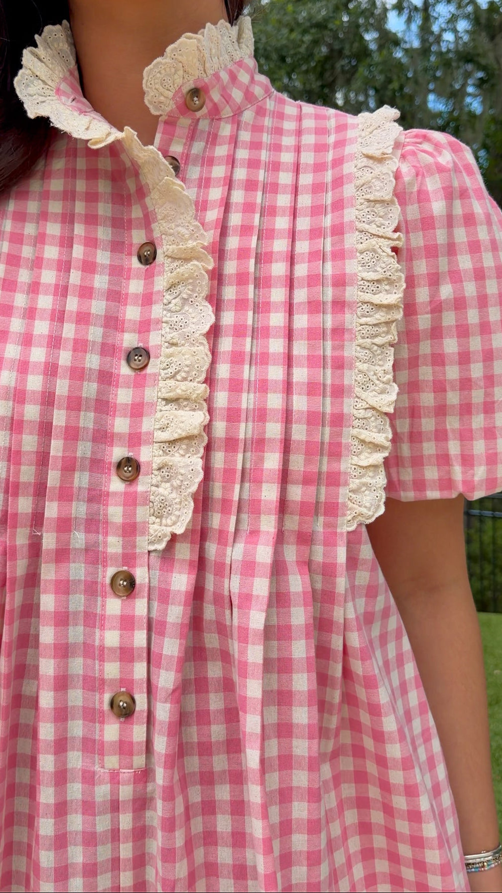 A close-up of a person wearing a pink and white gingham dress with cream lace trim and brown buttons, photographed outdoors with trees and grass in the background.