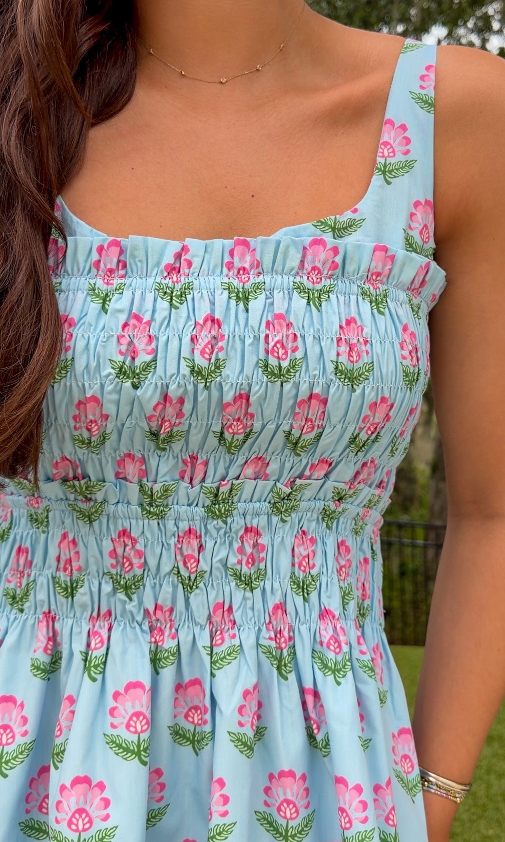 A close-up of a woman wearing a light blue smocked dress with a pink and green floral pattern, photographed outdoors with trees and grass in the background.