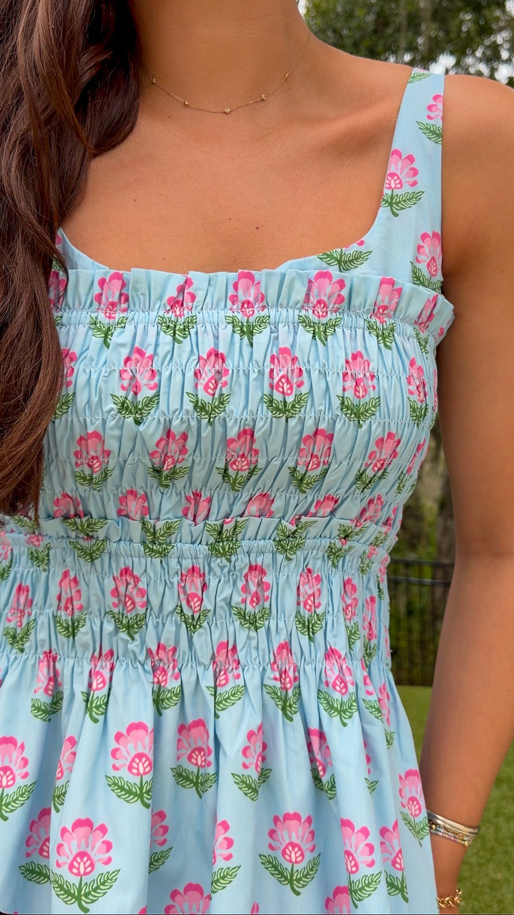 A close-up of a woman wearing a light blue smocked dress with a pink and green floral pattern, photographed outdoors with trees and grass in the background.
