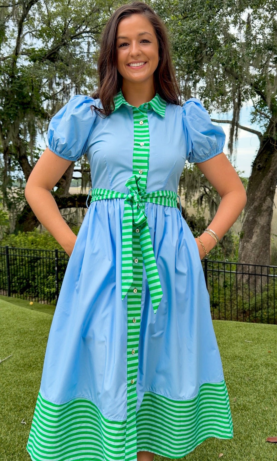 Sky blue midi dress featuring green striped contrast trim, a collared button-front design, puff sleeves, removable self-tie waist sash, side pockets, and a flowing midi-length skirt.