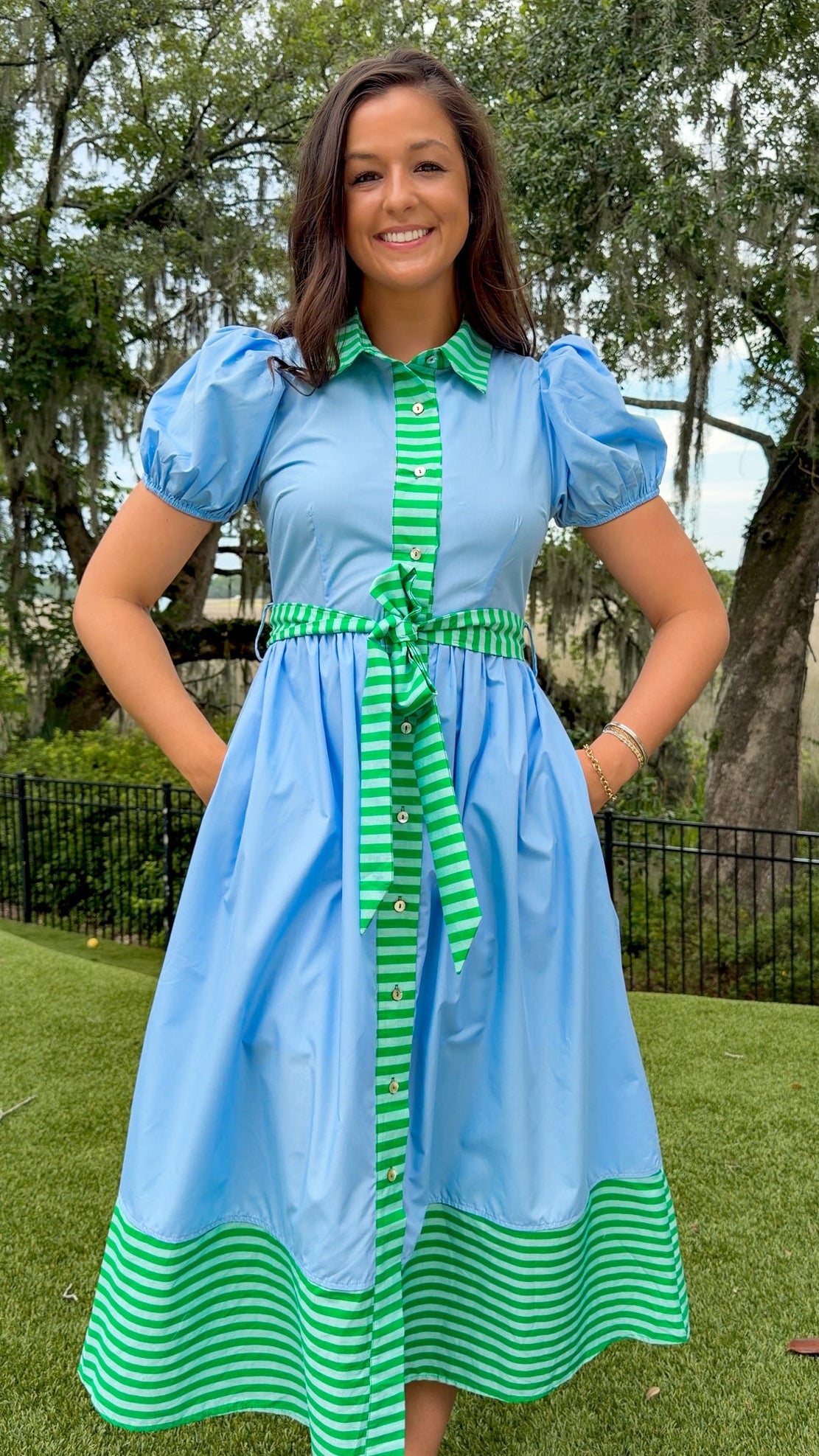 Sky blue midi dress featuring green striped contrast trim, a collared button-front design, puff sleeves, removable self-tie waist sash, side pockets, and a flowing midi-length skirt.