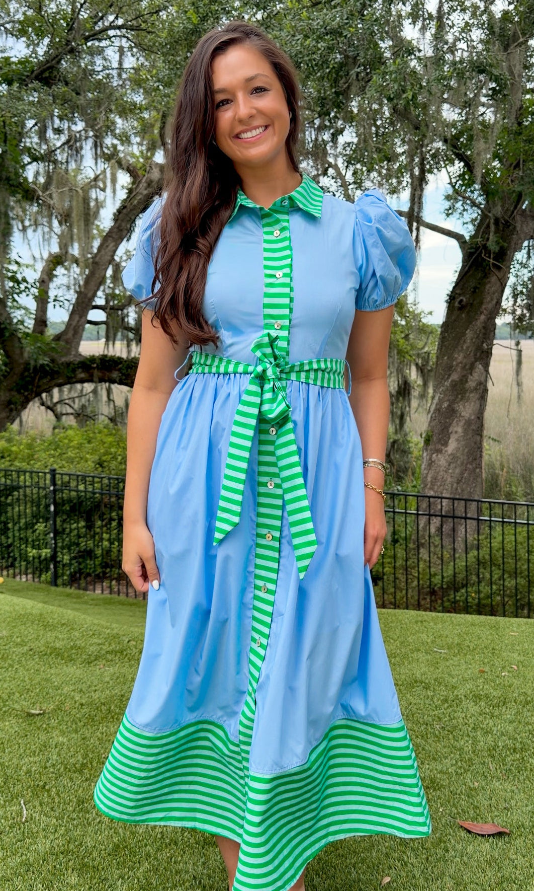 Sky blue midi dress featuring green striped contrast trim, a collared button-front design, puff sleeves, removable self-tie waist sash, side pockets, and a flowing midi-length skirt.