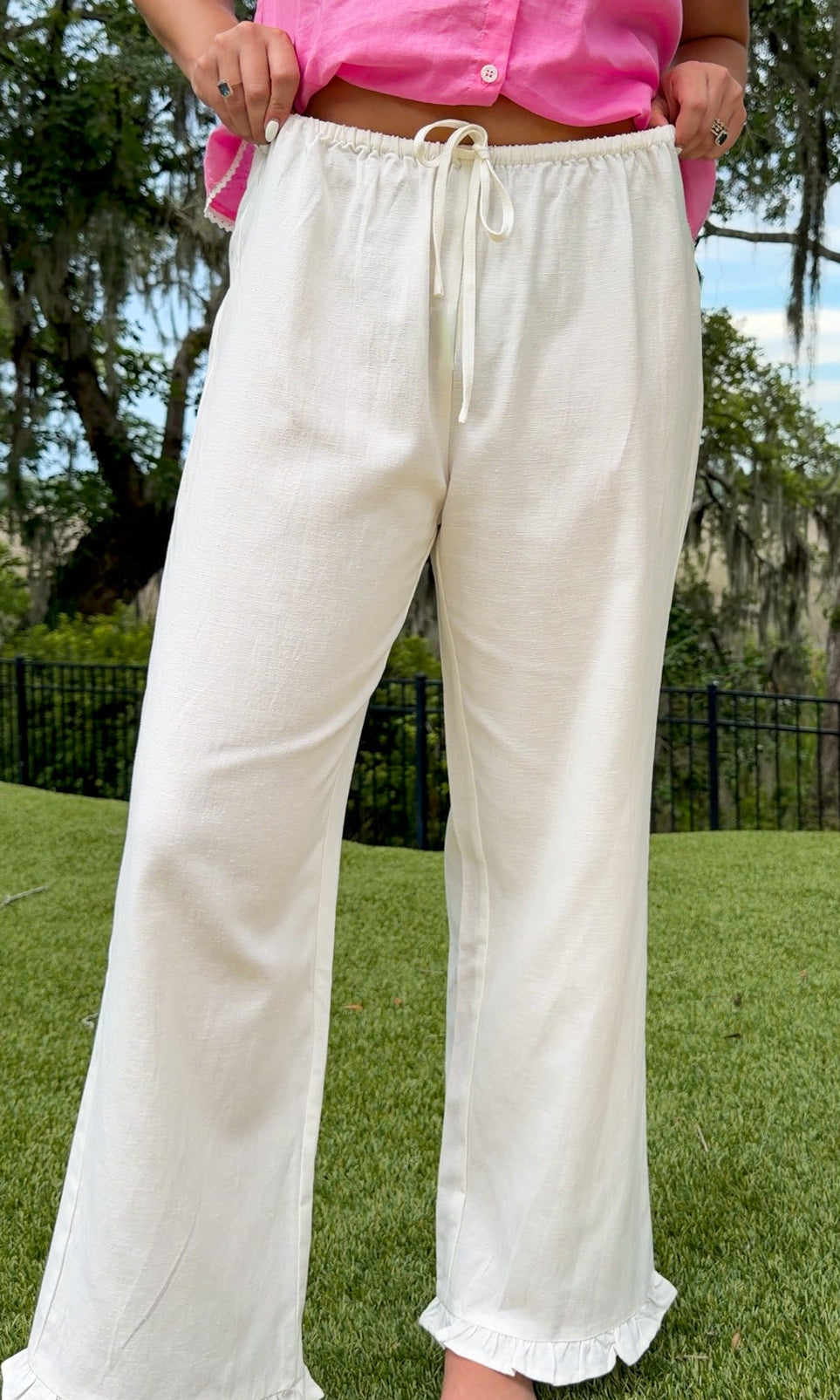 White full-length straight-leg pants featuring an elastic drawstring waistband, side and back patch pockets, delicate ruffle trim at the hem, and a relaxed, lightweight fit.