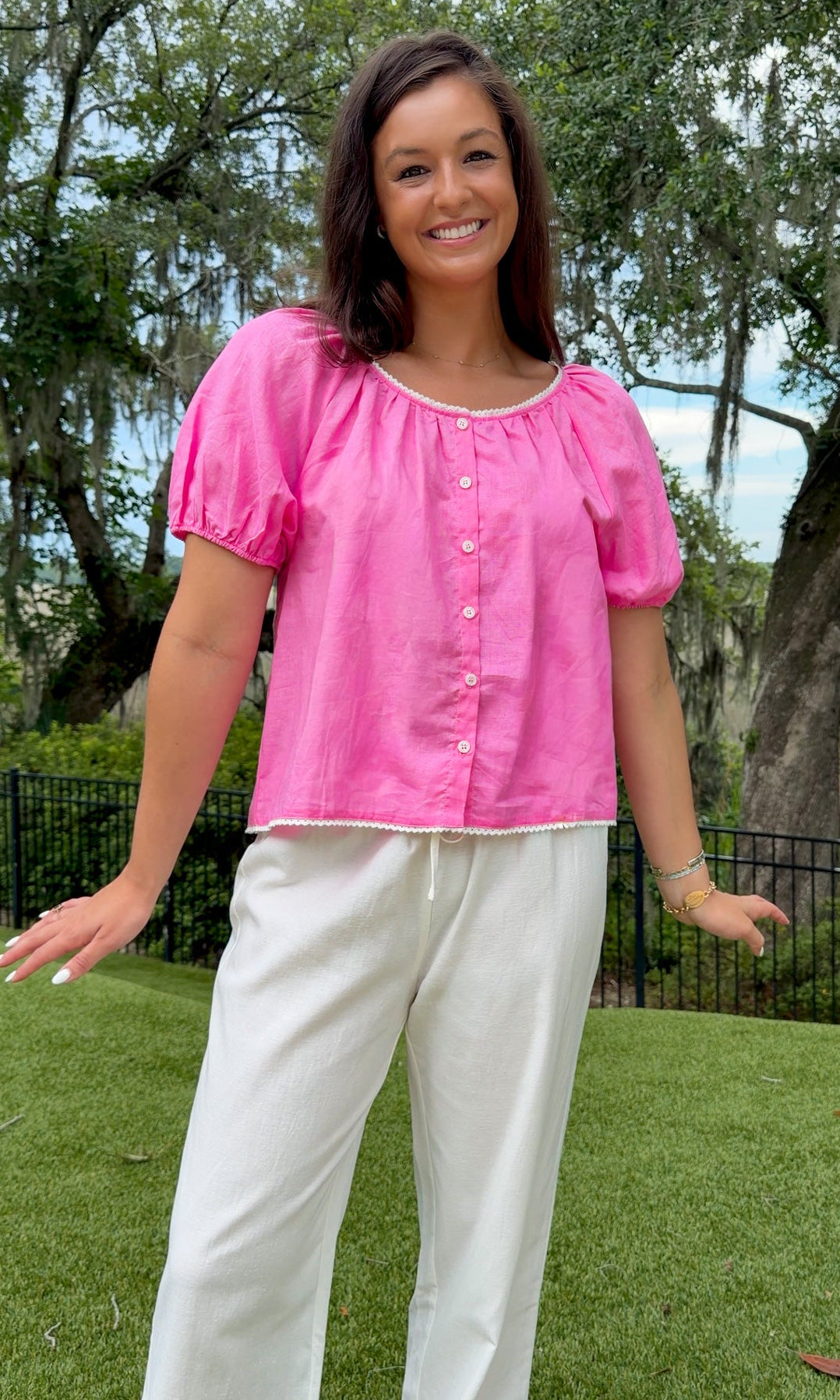 A woman wearing a bright pink short-sleeve puff-sleeve top with white buttons and white wide-leg pants, standing outdoors on a green lawn with trees in the background.