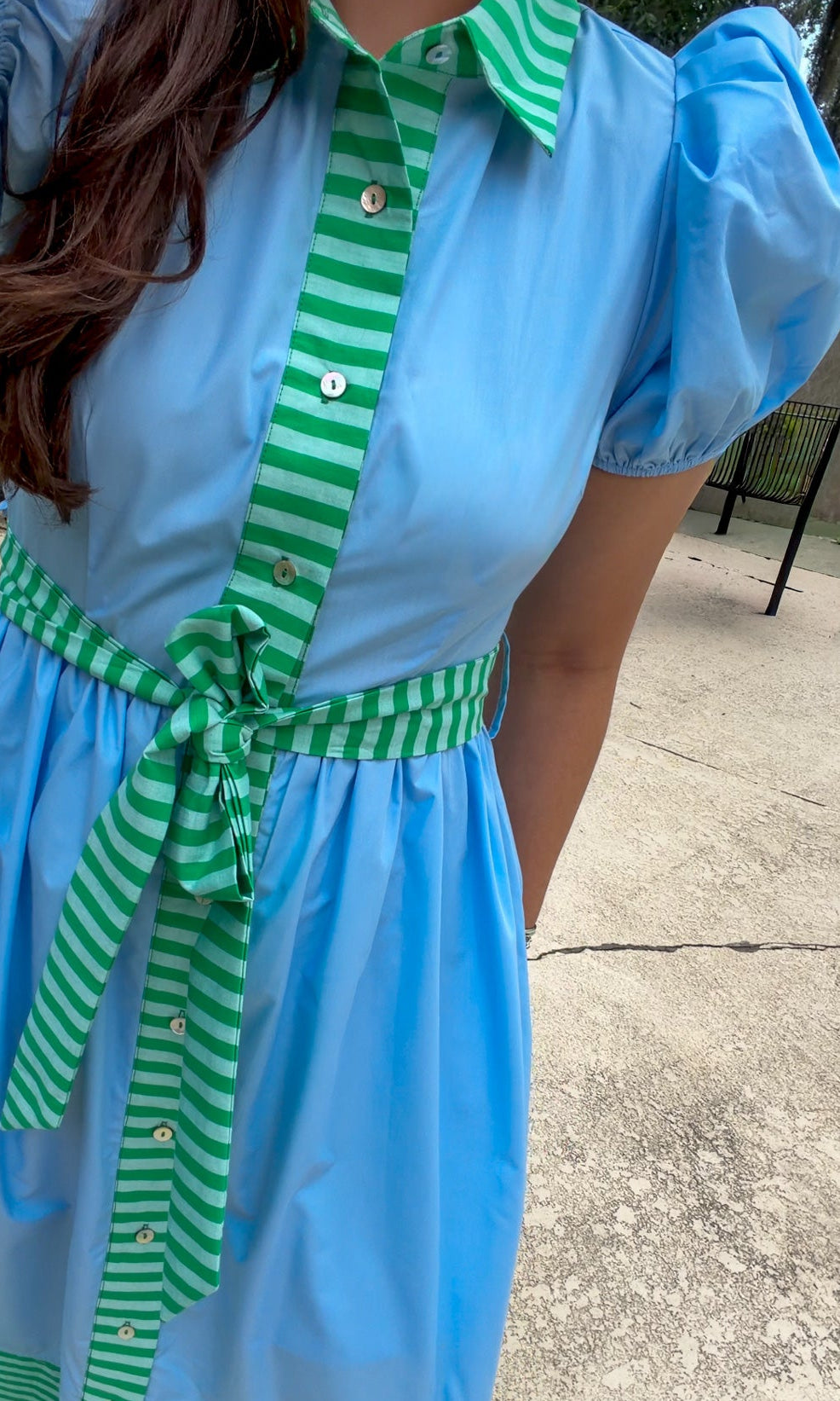 A woman wearing a light blue short-sleeve shirt dress with green and white striped trim on the collar, front placket, waist tie, and hem, standing outdoors on a concrete surface.