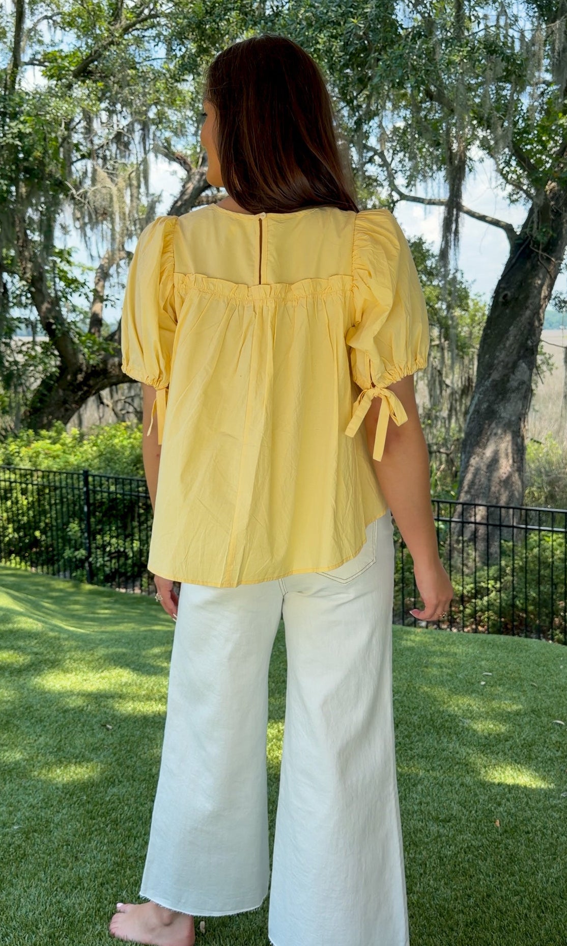 Butter yellow babydoll top featuring a round neckline, tiered shirring, short puff sleeves with adjustable tie cuffs, a ruffle-trimmed back, keyhole button closure, and a relaxed semi-cropped silhouette.
