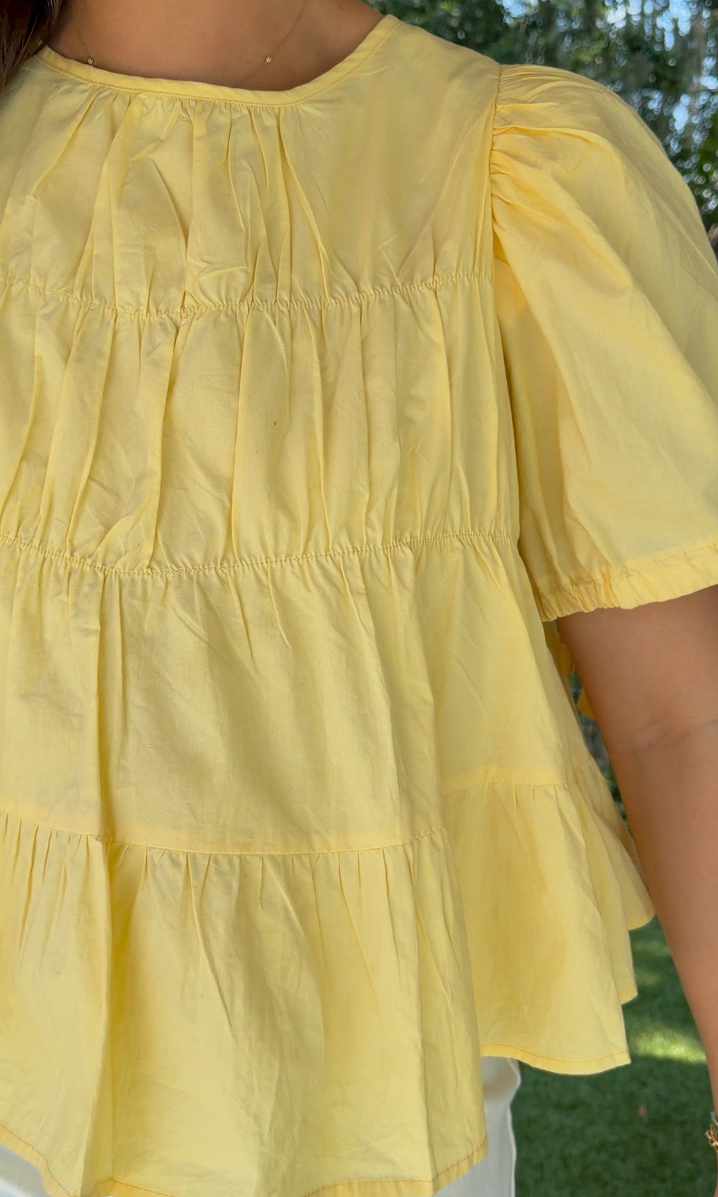Butter yellow babydoll top featuring a round neckline, tiered shirring, short puff sleeves with adjustable tie cuffs, a ruffle-trimmed back, keyhole button closure, and a relaxed semi-cropped silhouette.
