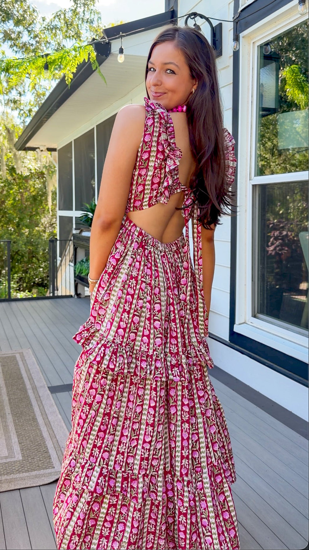 A woman wearing a pink and white patterned tiered maxi dress with a cutout back, standing on a wooden deck in front of a house.