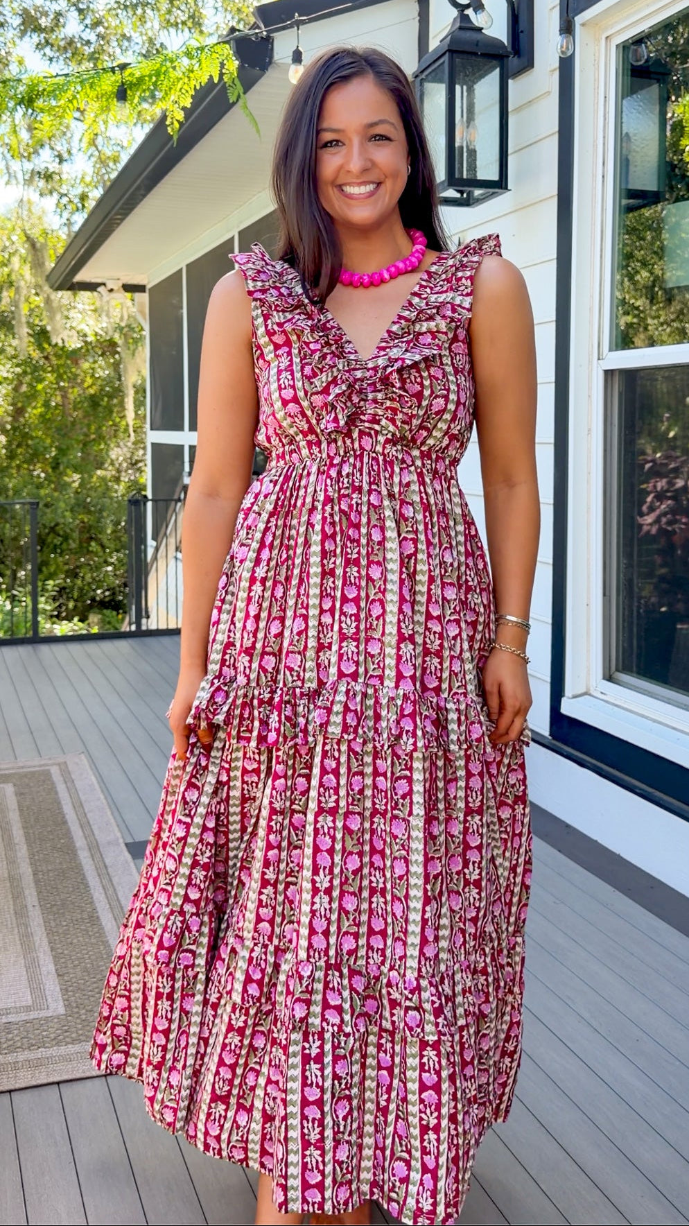 A woman stands on a wooden deck wearing a sleeveless tiered midi dress with a pink, red, and white floral pattern, paired with a bright pink necklace.