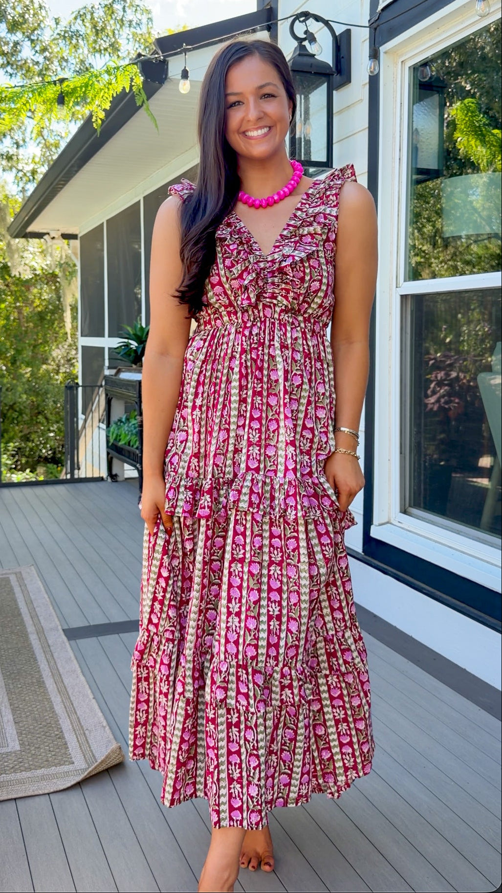 A woman stands on a wooden deck in front of a house, wearing a sleeveless tiered maxi dress with a pink, red, and beige floral pattern, paired with a bright pink necklace.