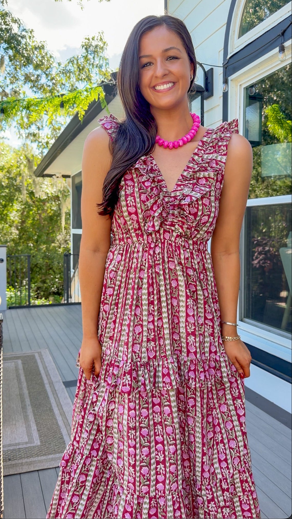 A woman wearing a sleeveless tiered maxi dress with a pink, red, and beige floral pattern, standing on a wooden deck in front of a house.