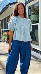 Woman wearing a light blue blouse with floral patterns and blue jeans standing outdoors.