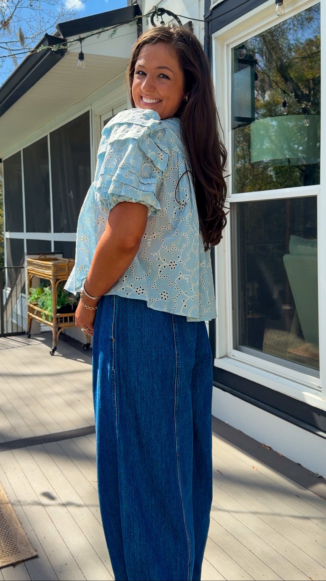 Woman wearing a light blue blouse with floral patterns and blue jeans standing outdoors.
