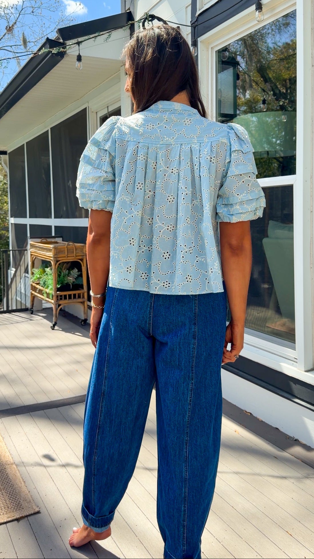 Woman wearing a light blue blouse with floral patterns and blue jeans standing outdoors.