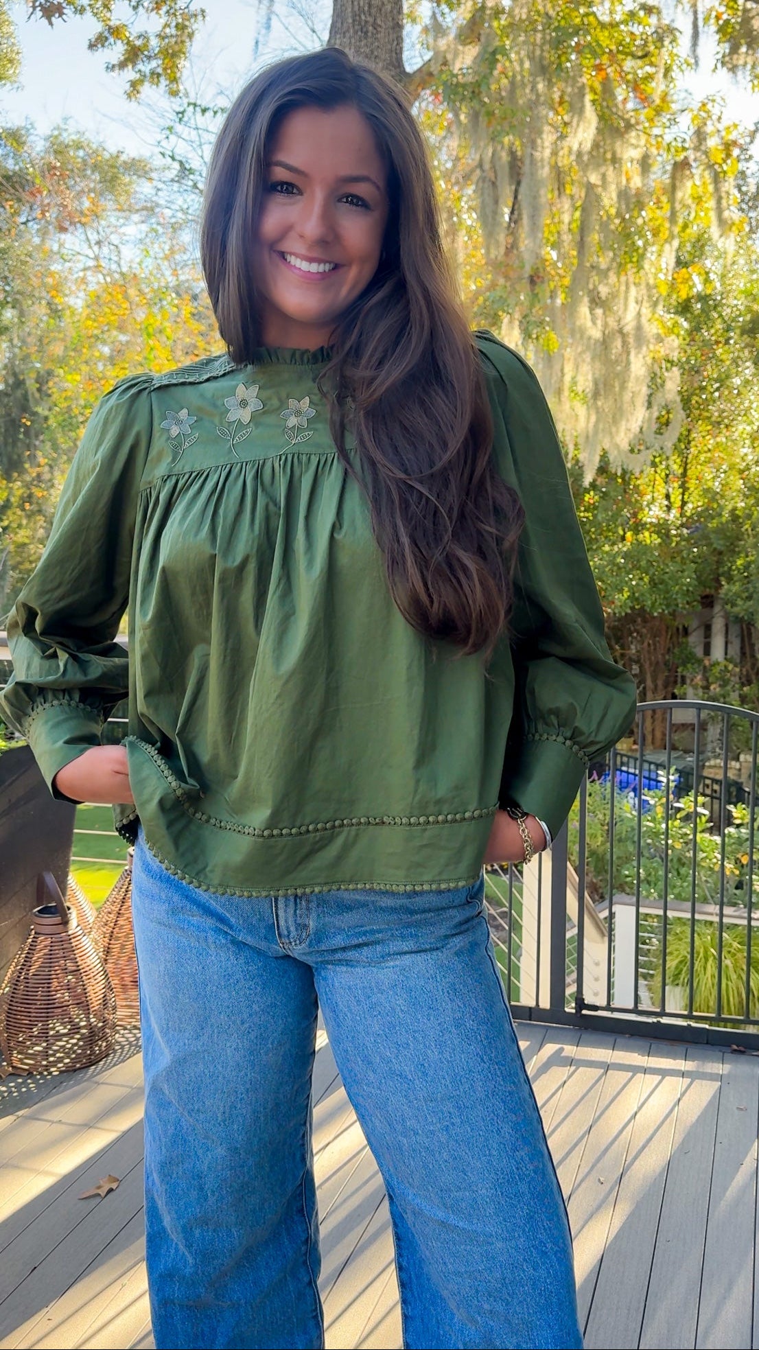 Woman wearing a green blouse and blue jeans standing on a wooden deck with trees in the background.