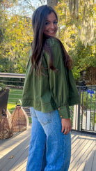 Woman wearing a green jacket and blue jeans standing on a wooden deck with trees in the background