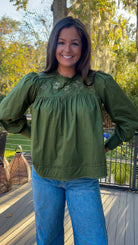Woman wearing a green blouse with floral embroidery and blue jeans outdoors.