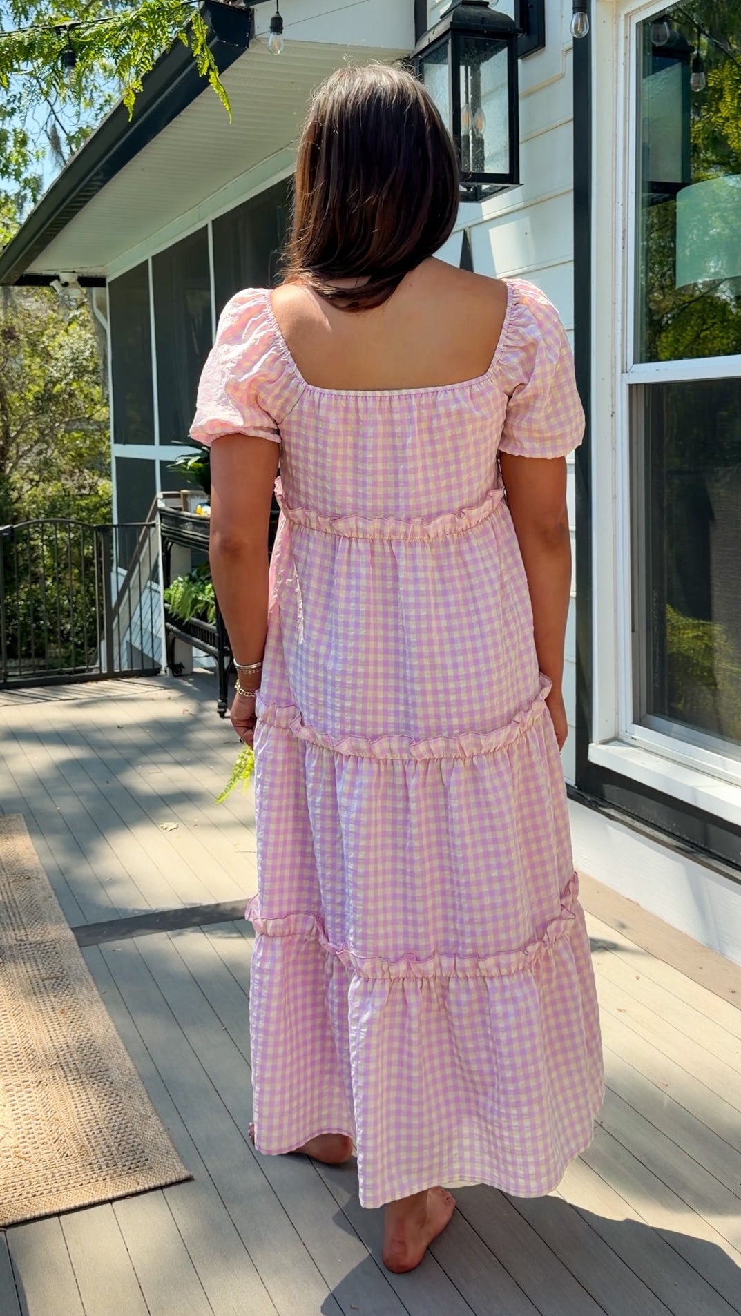 Woman in a pink checkered dress standing on a wooden deck with a house in the background.