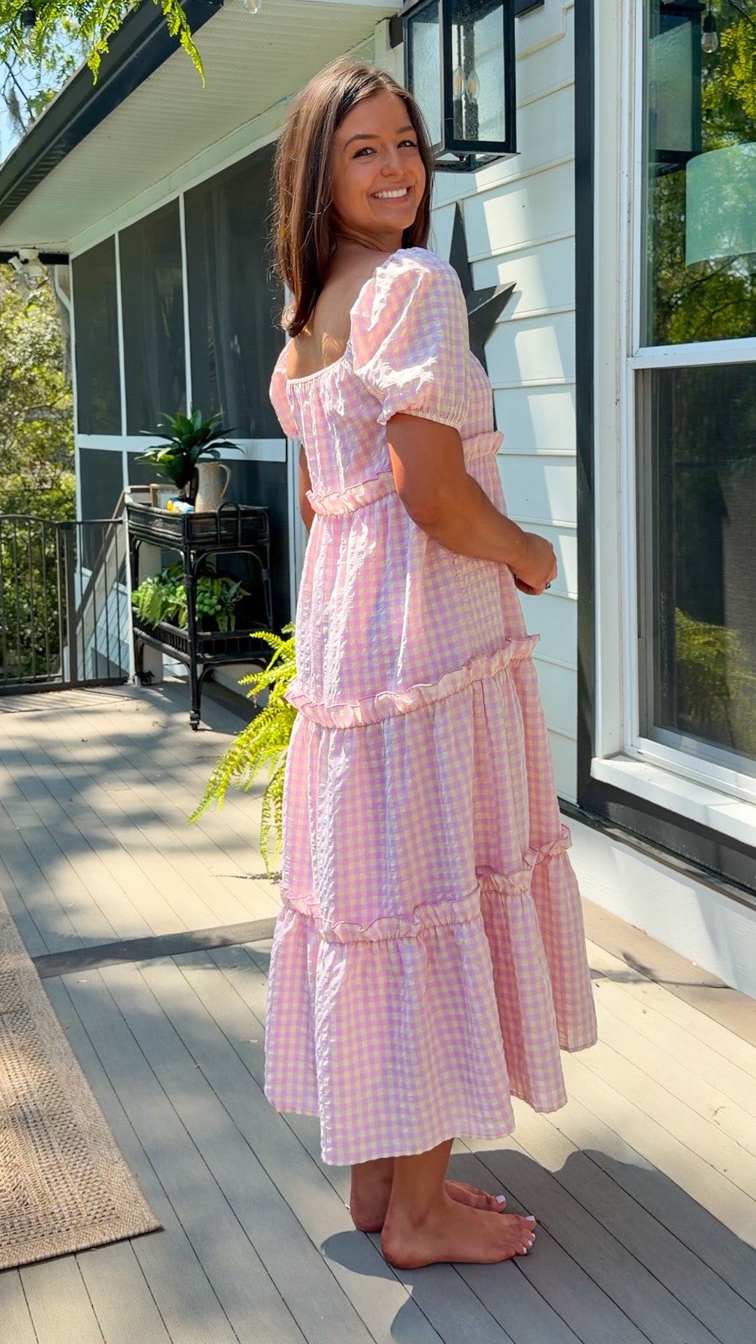 Woman in a pink checkered dress standing on a deck with a house in the background