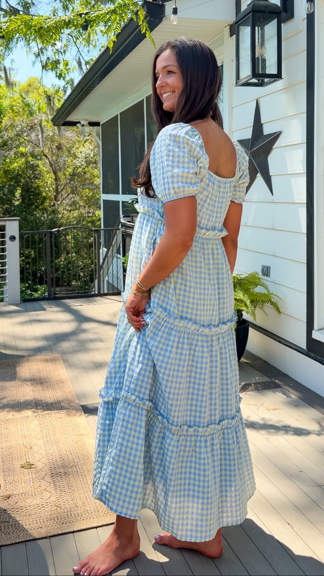 Woman in a blue checkered dress standing on a wooden deck with a house in the background.