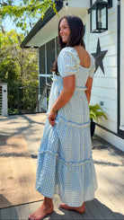 Woman in a blue checkered dress standing on a wooden deck with a house in the background.