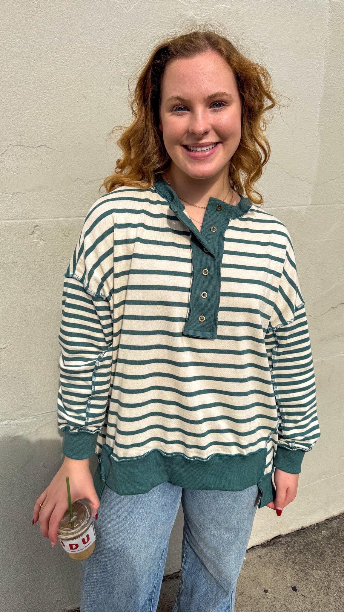 Woman wearing a striped shirt and jeans, holding a drink against a plain wall.