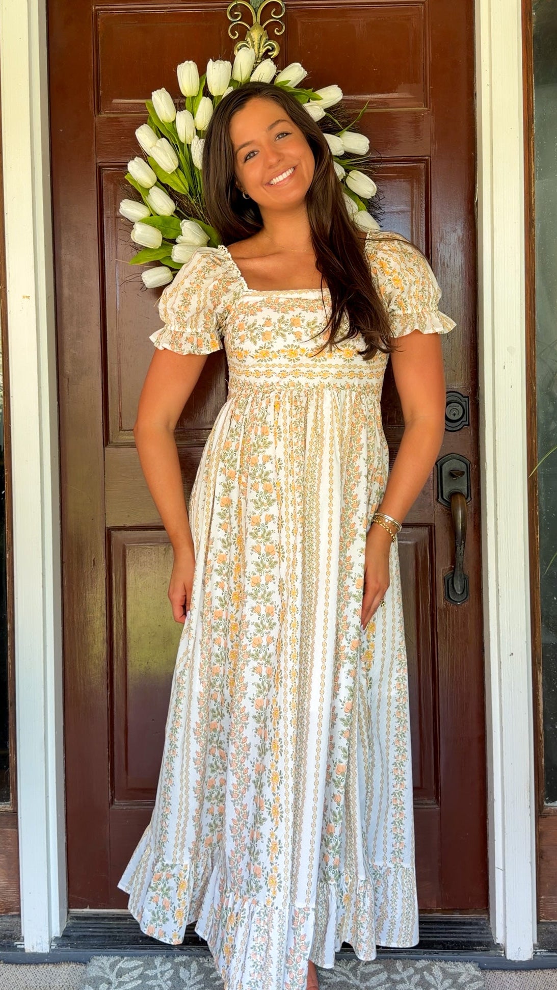 Woman in a floral dress standing in front of a door with a wreath, smiling.