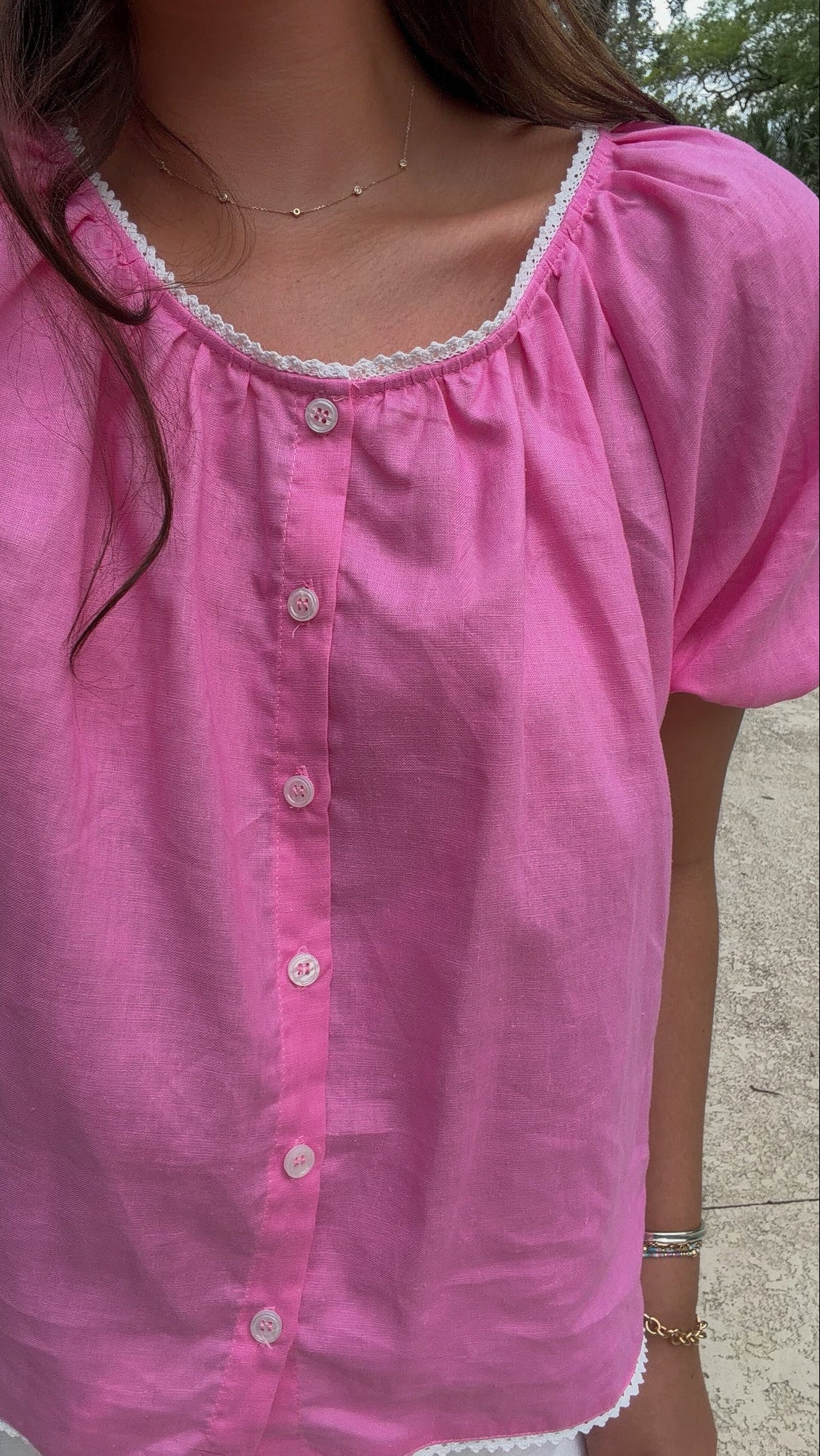A close-up of a woman wearing a pink short-sleeve button-front blouse with white lace trim around the neckline and hem.