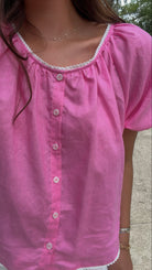 A close-up of a woman wearing a pink short-sleeve button-front blouse with white lace trim around the neckline and hem.