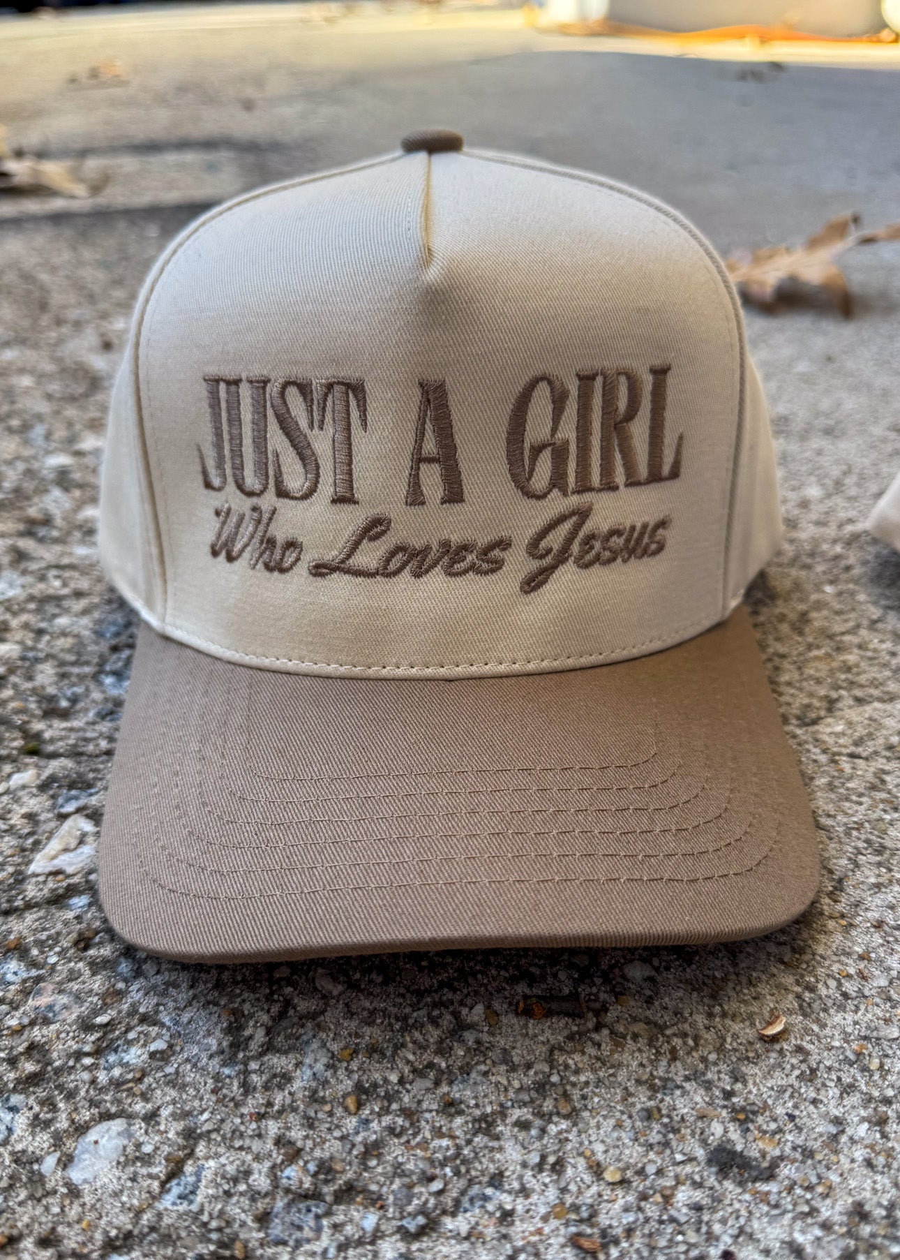 Beige cap with 'Just a Girl Who Loves Jesus' text on a concrete surface