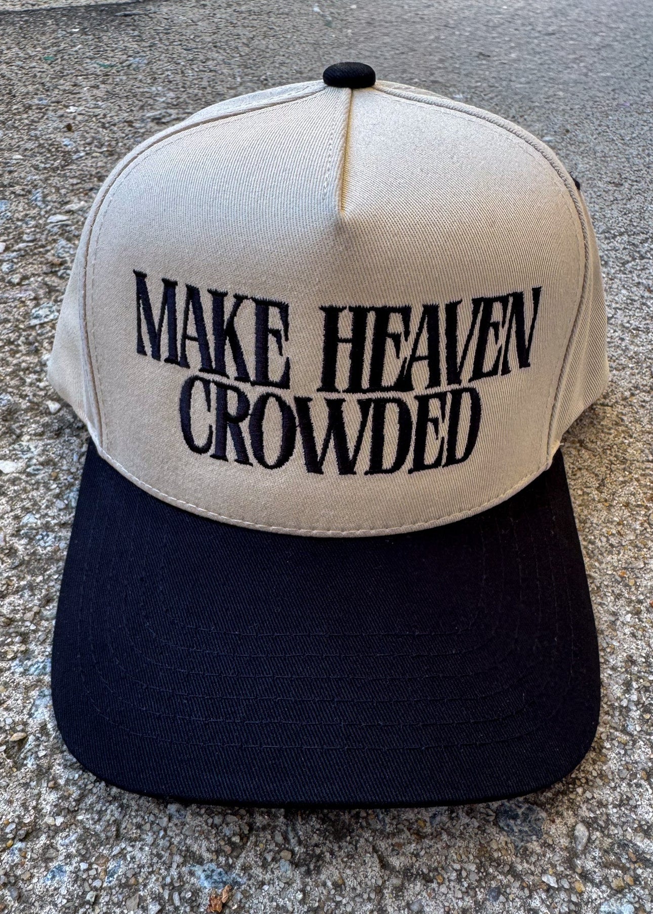 Beige and black cap with 'MAKE HEAVEN CROWDED' text on a concrete surface