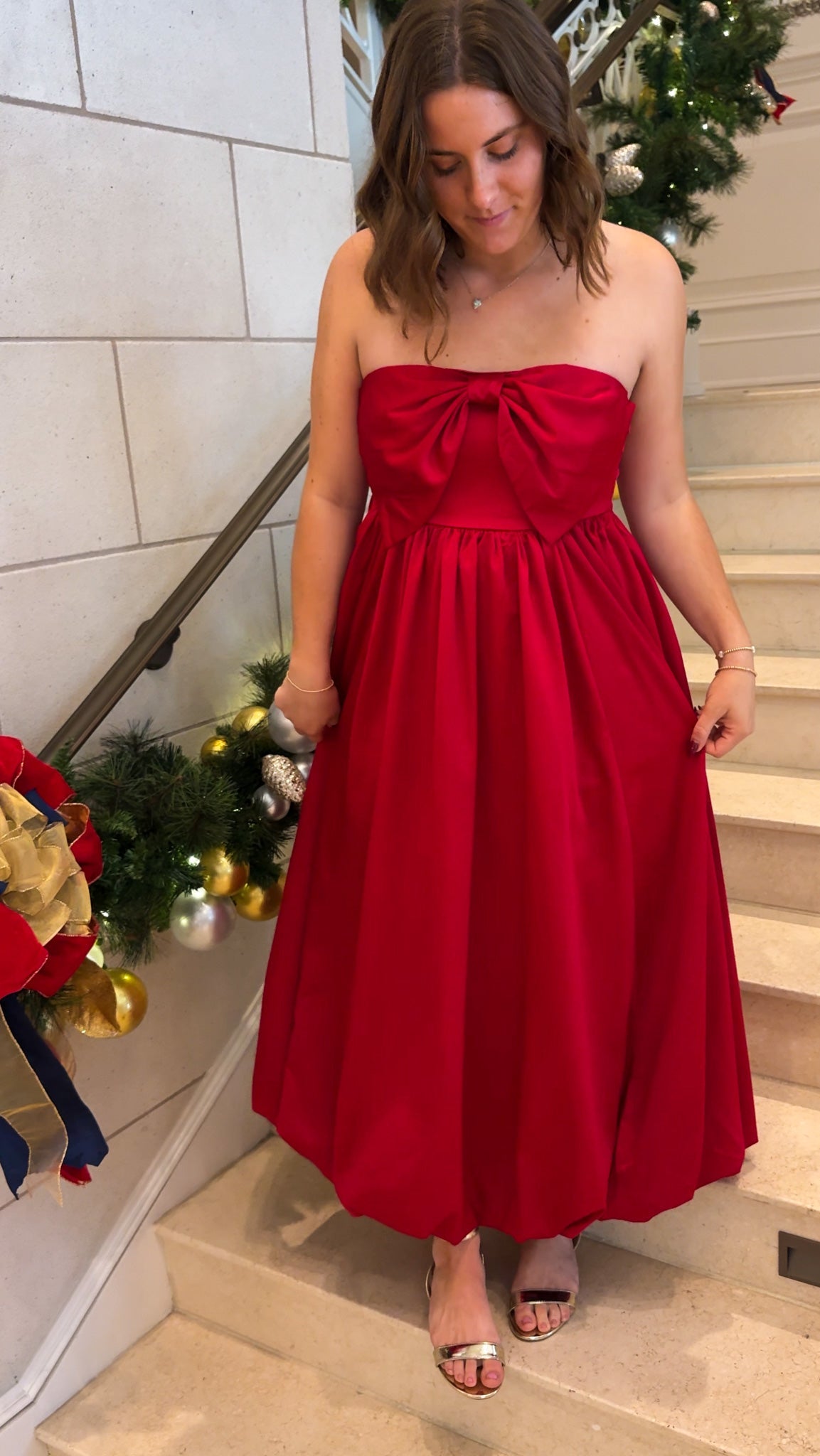 Red strapless midi dress with large front bow detail, full gathered skirt, and smocked back panel, worn on staircase with holiday decor.