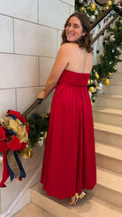 Red strapless midi dress with large front bow detail, full gathered skirt, and smocked back panel, worn on staircase with holiday decor.