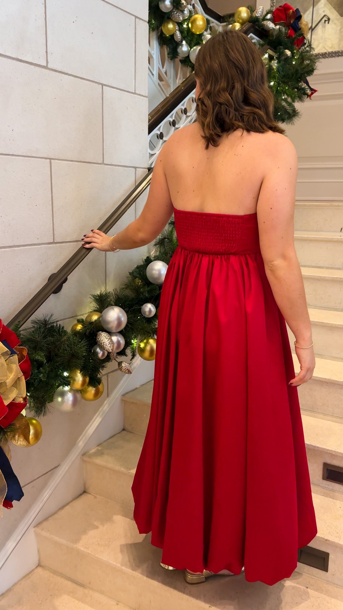 Red strapless midi dress with large front bow detail, full gathered skirt, and smocked back panel, worn on staircase with holiday decor.