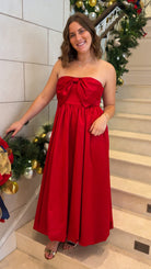 Red strapless midi dress with large front bow detail, full gathered skirt, and smocked back panel, worn on staircase with holiday decor.
