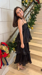 Black tiered midi dress with spaghetti straps, sweetheart-style neckline, and back zipper closure, worn on indoor staircase with holiday decor.