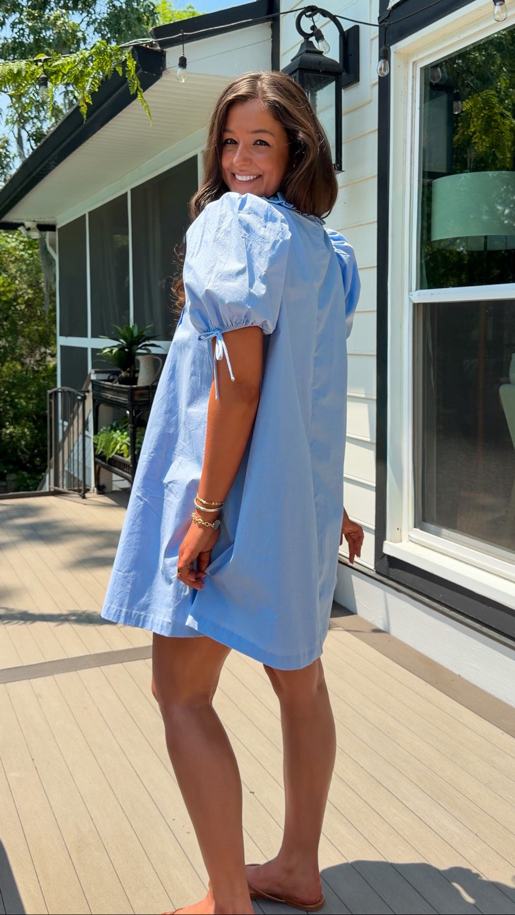 Woman wearing a light blue dress standing on a wooden deck with a house in the background.