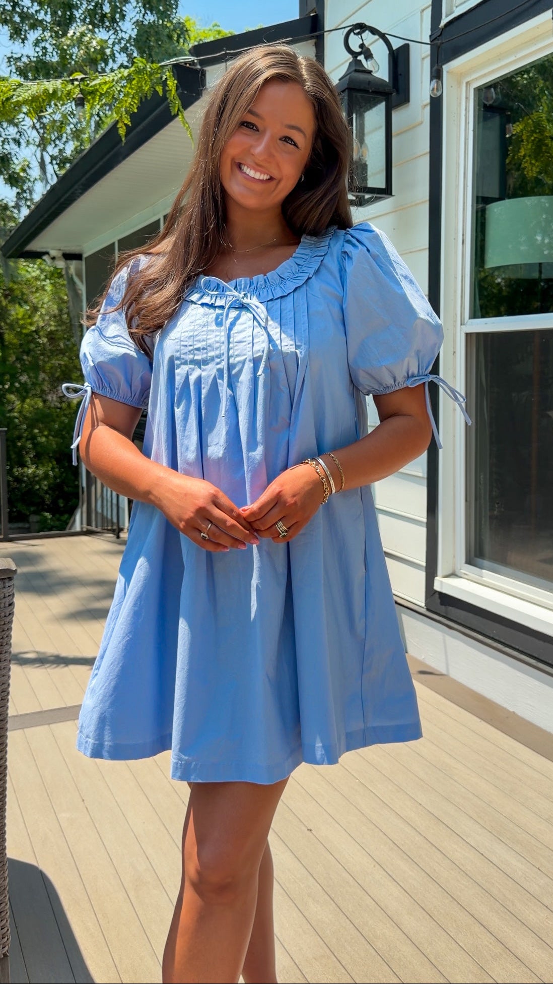 Woman in a light blue dress standing on a wooden deck with a house in the background.