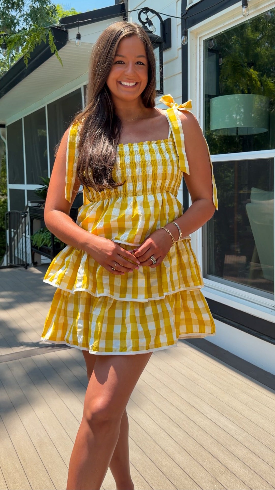 Woman in a yellow checkered dress standing on a wooden deck with a house in the background.