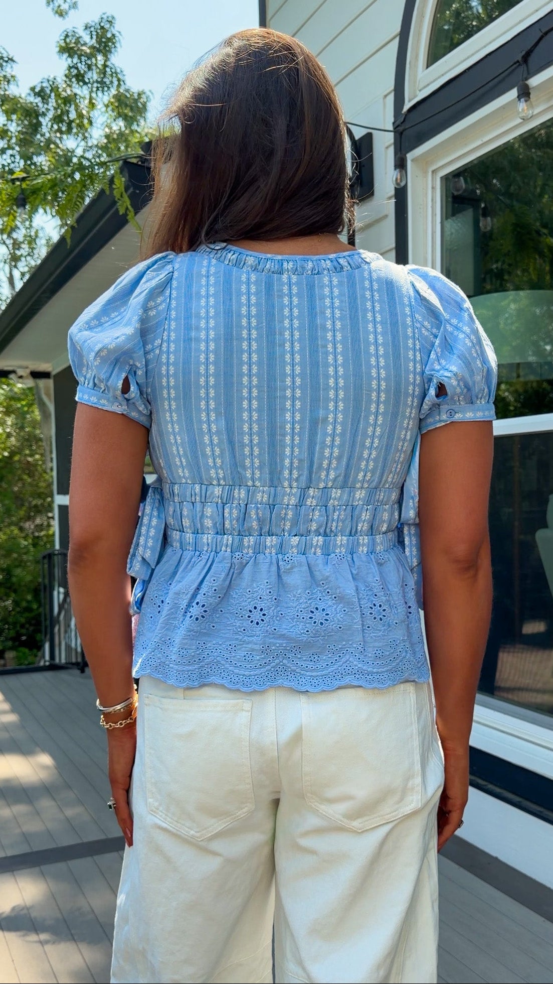 Person wearing a blue patterned top and beige pants standing outside a house.