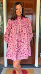 Model wearing a mauve floral corduroy babydoll dress with smocked bodice and velvet collar and cuffs standing in front of a wooden door.