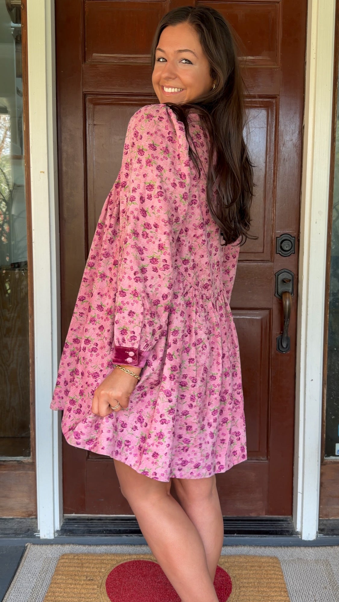 Model wearing a mauve floral corduroy babydoll dress with smocked bodice and velvet collar and cuffs standing in front of a wooden door.