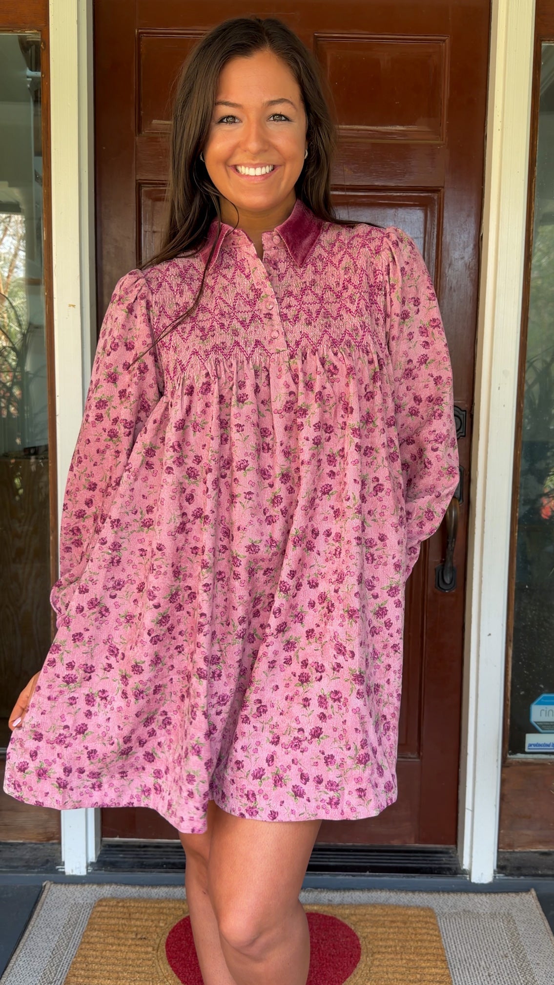 Model wearing a mauve floral corduroy babydoll dress with smocked bodice and velvet collar and cuffs standing in front of a wooden door.