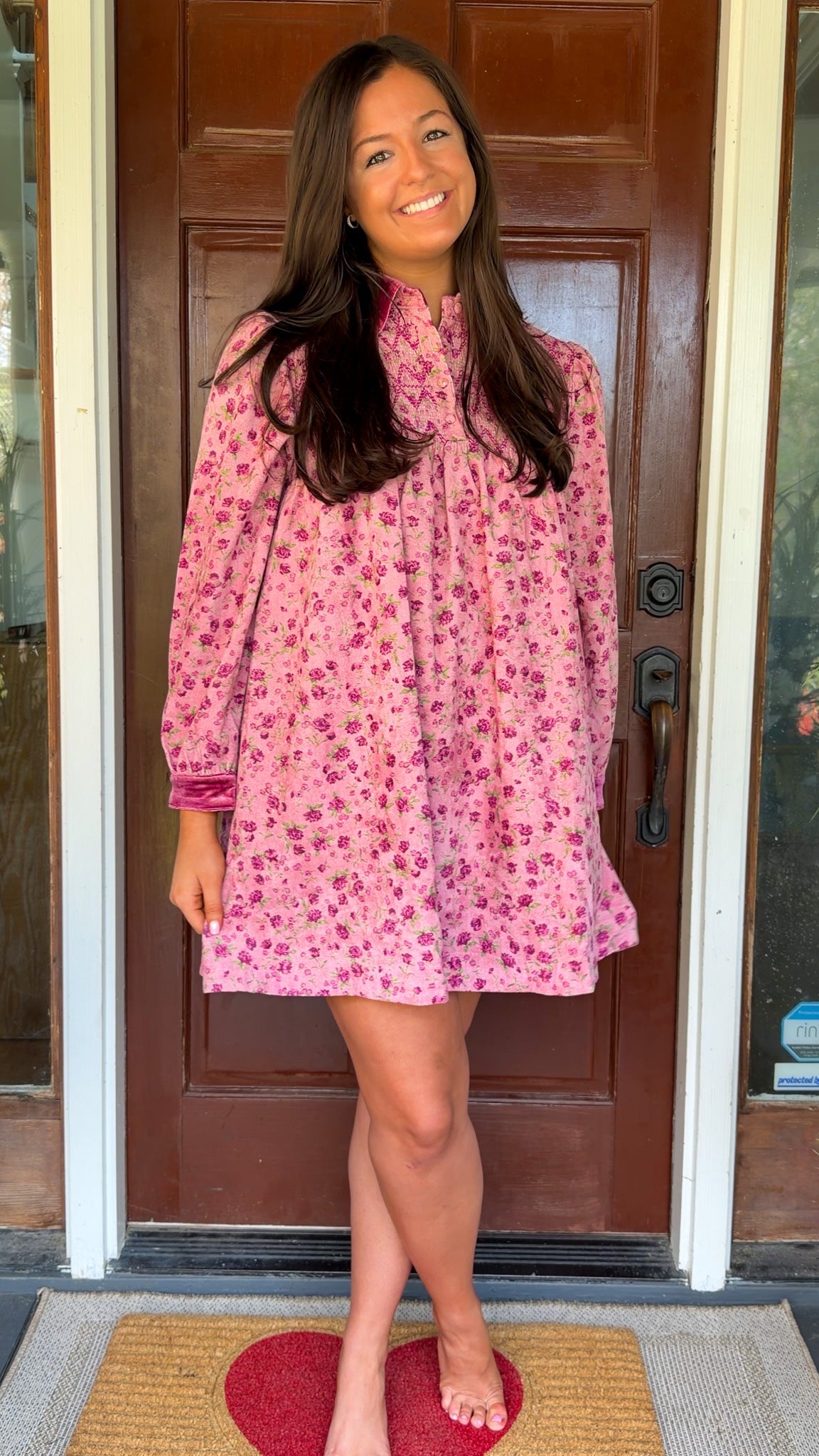 Model wearing a mauve floral corduroy babydoll dress with smocked bodice and velvet collar and cuffs standing in front of a wooden door.