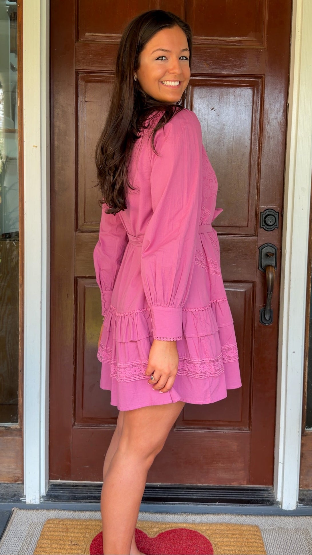 Mauve high-neck lace trim mini dress with long sleeves, self-tie waist, and tiered ruffle skirt worn by model standing in front of a wooden door
