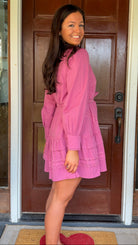 Mauve high-neck lace trim mini dress with long sleeves, self-tie waist, and tiered ruffle skirt worn by model standing in front of a wooden door